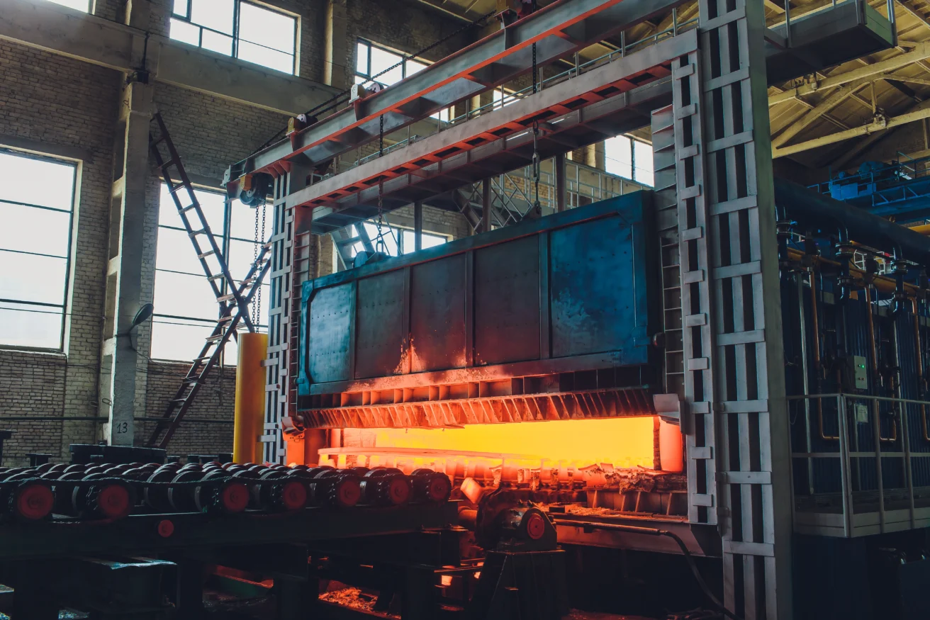 A large industrial furnace with a bright orange glow processes metal in a factory setting with high ceilings and large windows.