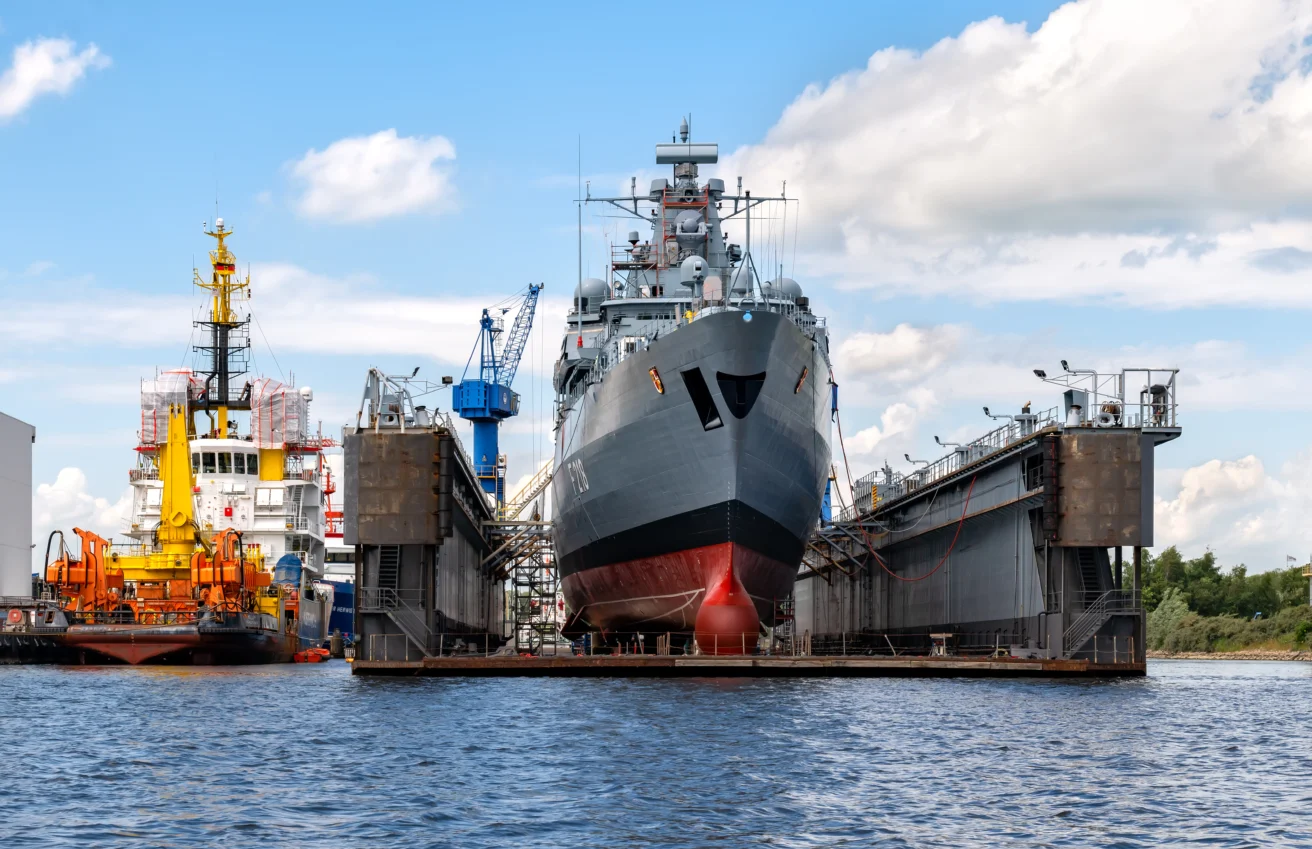 A large navy ship is docked in a floating dry dock for maintenance, with cranes and another vessel visible in the background.