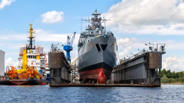 A large navy ship is docked in a floating dry dock for maintenance, with cranes and another vessel visible in the background.