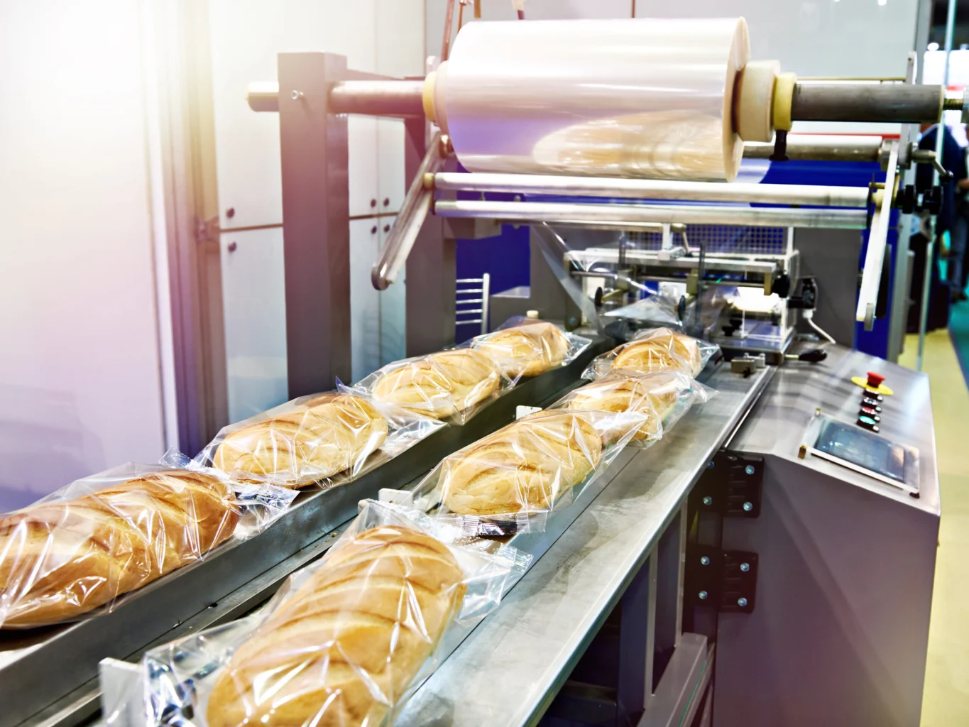 A bread packaging machine wrapping loaves in plastic on an assembly line in a factory setting.