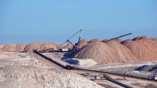 Mining site with large piles of minerals and machinery under a clear blue sky. Conveyor belts and heavy equipment are visible amid the mounds of earth and rocks.