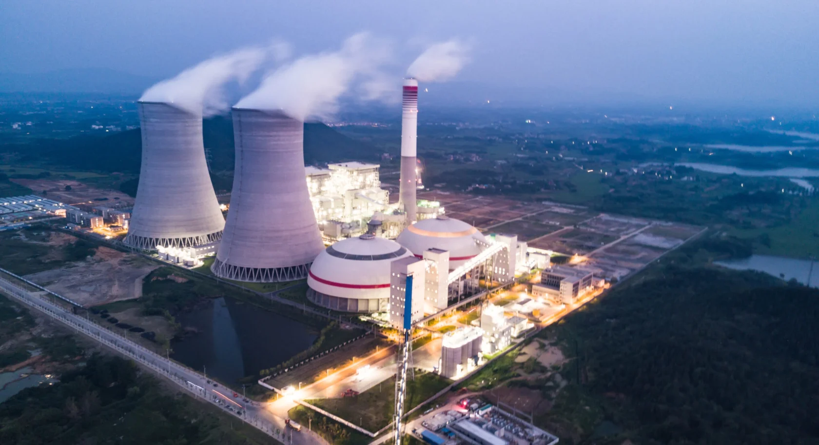 Aerial view of a power plant with two cooling towers emitting steam, surrounded by illuminated buildings and landscape at dusk.
