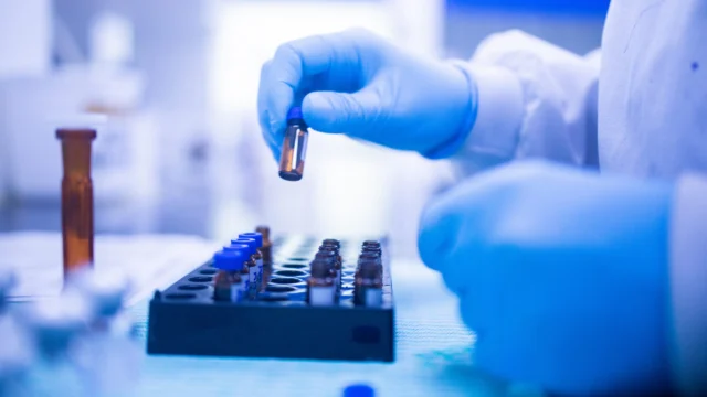 A gloved hand holds a small vial over a tray of test tubes in a laboratory setting.
