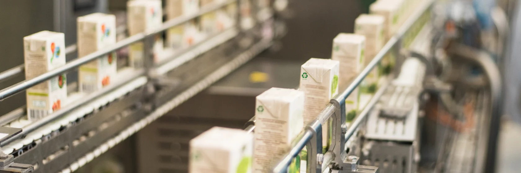 Rows of boxed beverages move along a conveyor belt in a factory setting, indicating an automated packaging or production line.