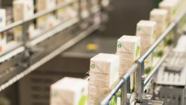 Rows of boxed beverages move along a conveyor belt in a factory setting, indicating an automated packaging or production line.