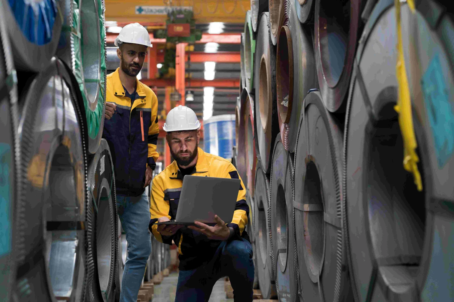 Team of male factory worker using laptop computer inspecting quality rolls of galvanized or metal sheet in in aluminum material warehouse