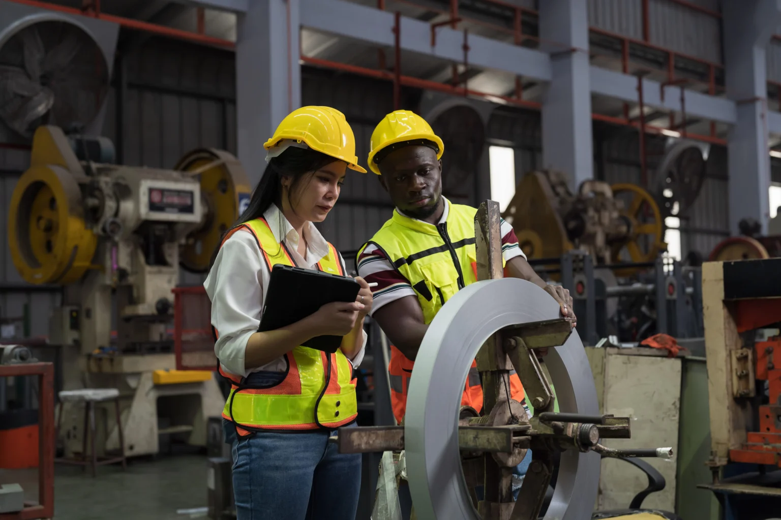 Two workers in safety gear inspect a metal roll in a factory. One holds a tablet, while the other examines the roll on machinery.