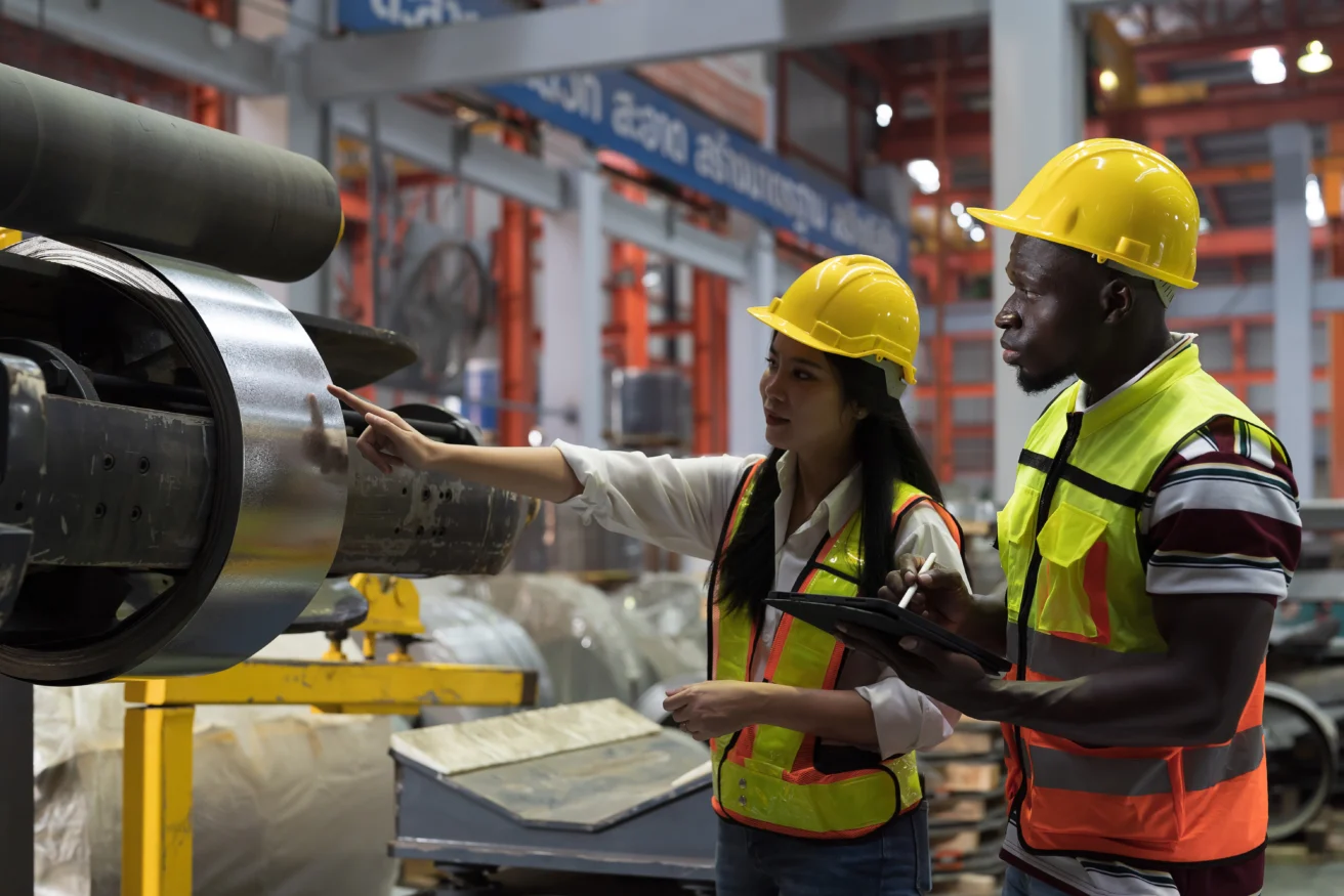 Two factory workers wearing safety hats and vests inspect machinery in an industrial setting; one points at equipment while the other takes notes on a clipboard.