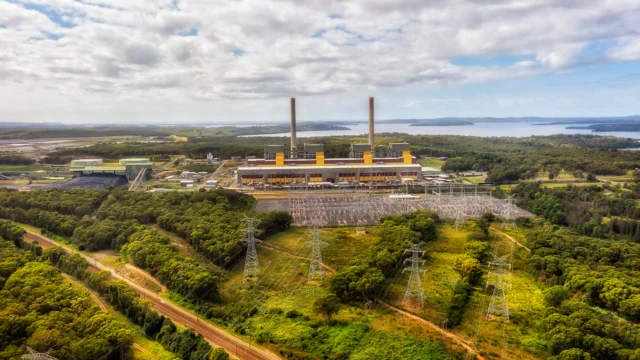 Aerial view of a large power plant with two tall chimneys, surrounded by trees, power lines, and a body of water in the background under a partly cloudy sky.