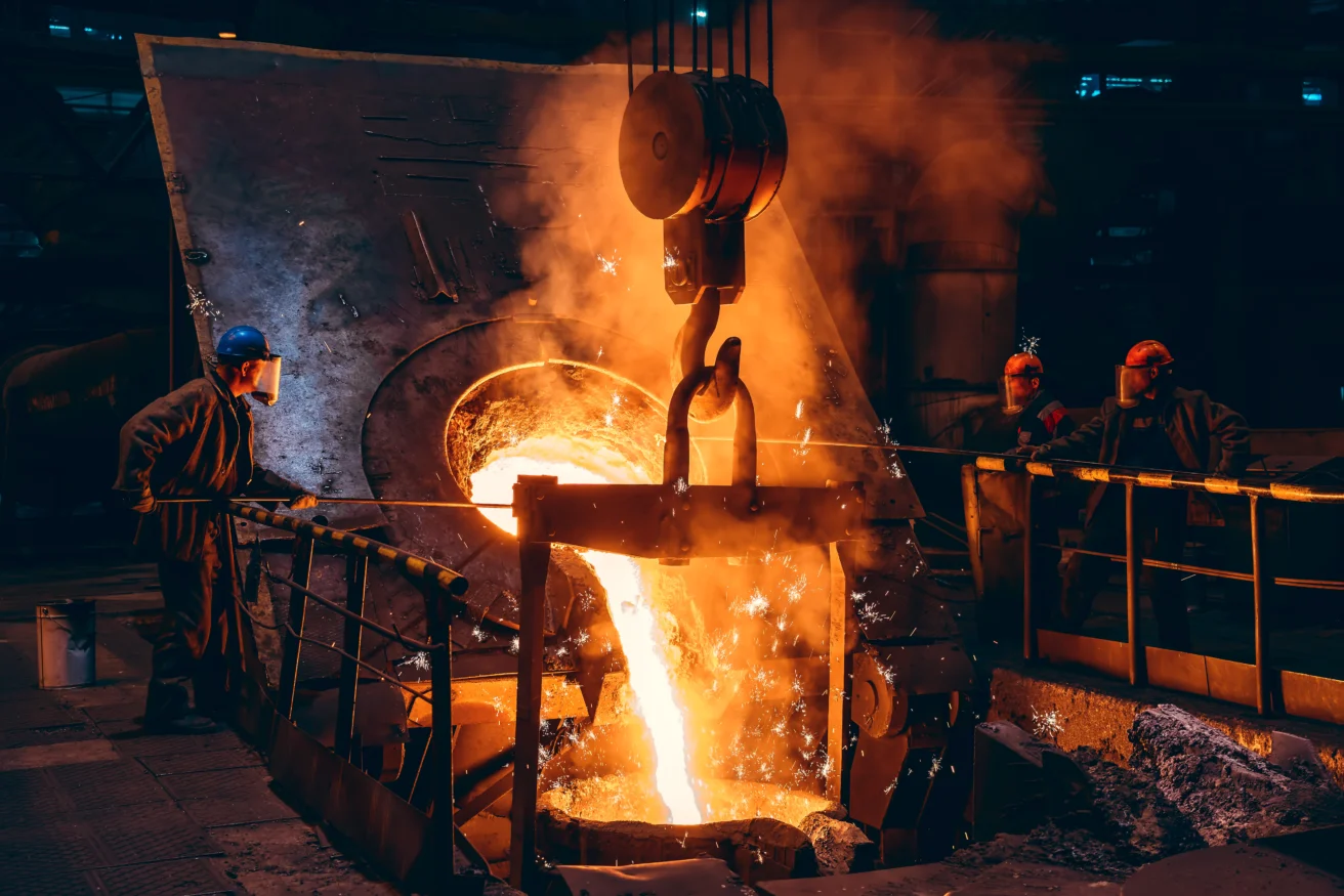 Industrial scene with workers in protective gear overseeing molten metal being poured from a large container in a foundry, with bright orange light and steam.