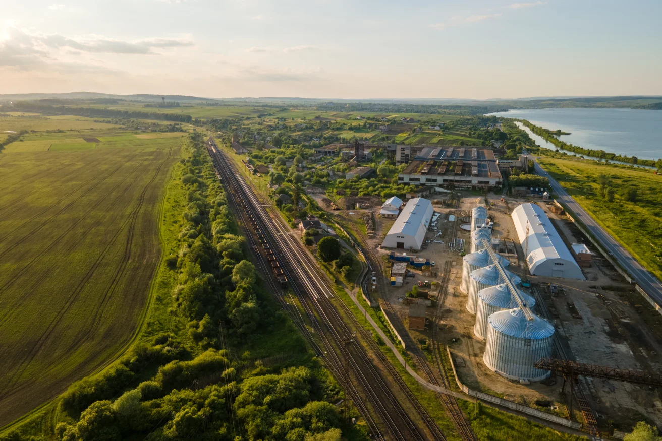 Aerial view of a rural industrial complex with silos, warehouses, nearby railway tracks, farmland, and a river or lake on the right.