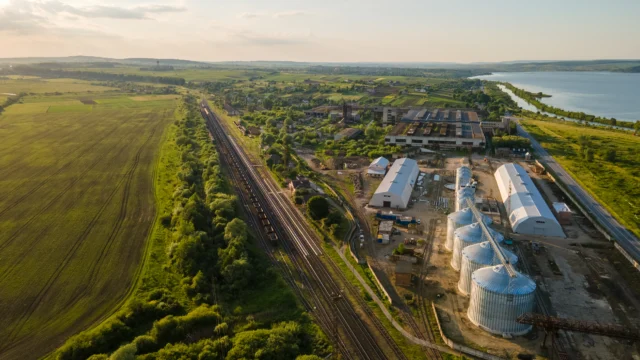 Aerial view of a rural industrial complex with silos, warehouses, nearby railway tracks, farmland, and a river or lake on the right.
