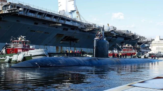 A submarine is docked by a large ship with two tugboats nearby, against a clear sky.