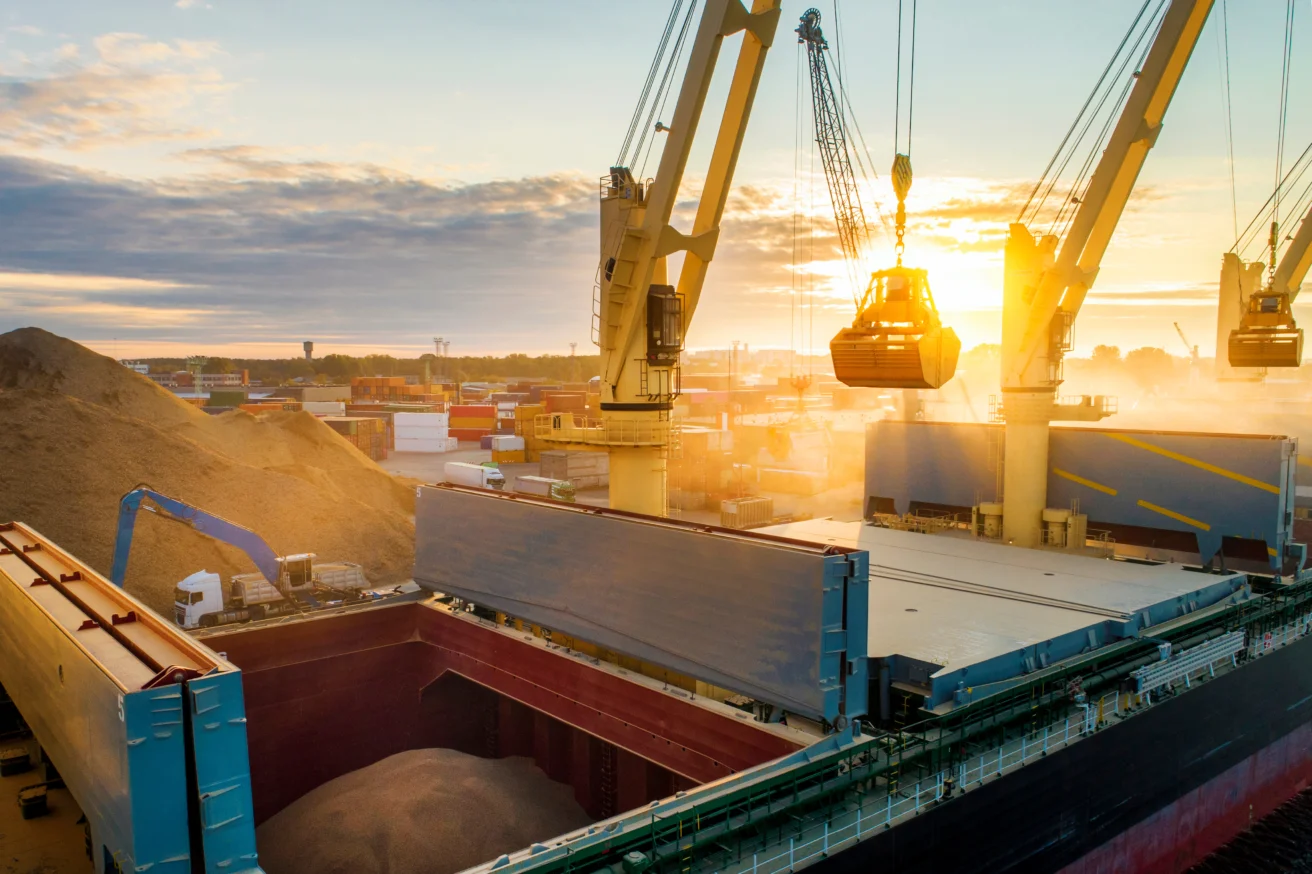 A cargo ship is being loaded with bulk material by cranes at an industrial port during sunset; shipping containers and machinery are visible in the background.