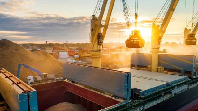 A cargo ship is being loaded with bulk material by cranes at an industrial port during sunset; shipping containers and machinery are visible in the background.