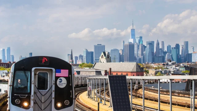 A New York City subway F train travels on elevated tracks with the Manhattan skyline and One World Trade Center visible in the background.
