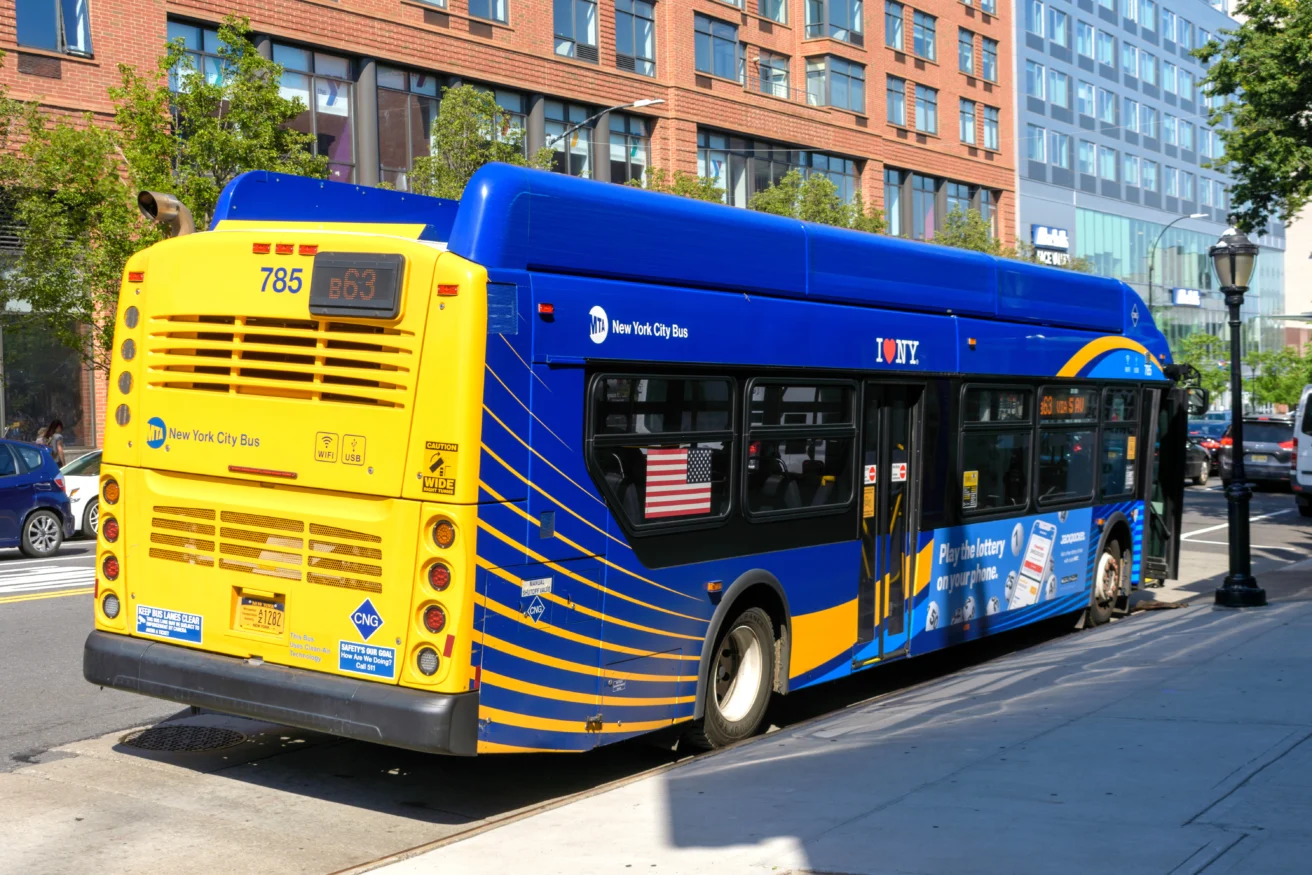 A New York City MTA bus, route 863, is parked at a curb near a sidewalk and brick apartment buildings on a sunny day.