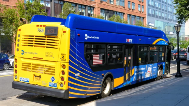 A New York City MTA bus, route 863, is parked at a curb near a sidewalk and brick apartment buildings on a sunny day.