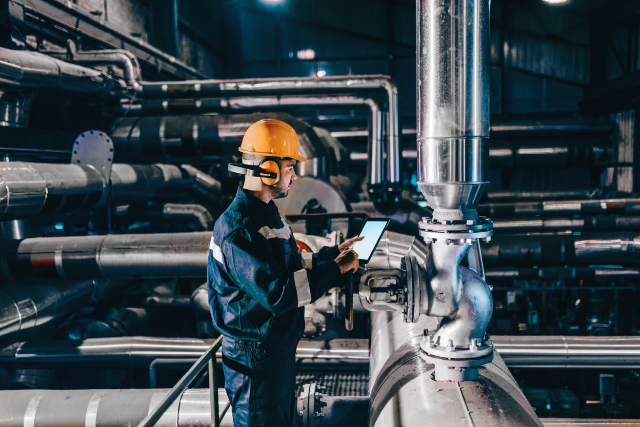 Person wearing a hard hat and protective gear inspects industrial piping with a tablet inside a large facility or factory.