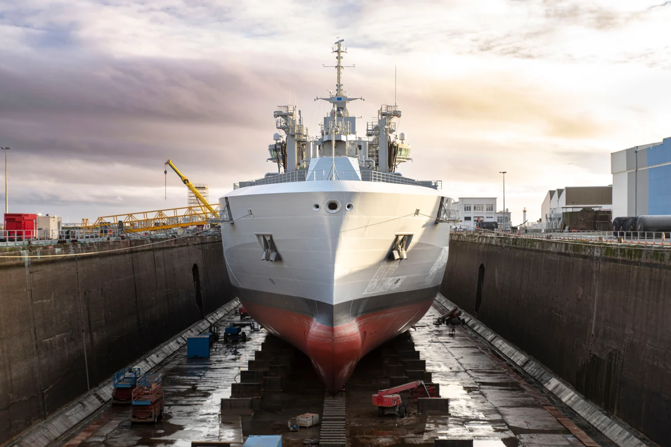 A large naval ship is positioned in a dry dock for maintenance, with industrial equipment and buildings visible in the background.