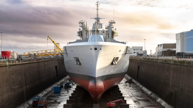 A large naval ship is positioned in a dry dock for maintenance, with industrial equipment and buildings visible in the background.