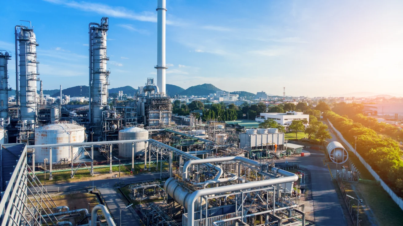 Industrial refinery complex with tall distillation towers, storage tanks, and pipelines set against a cityscape and mountains under a clear sky.