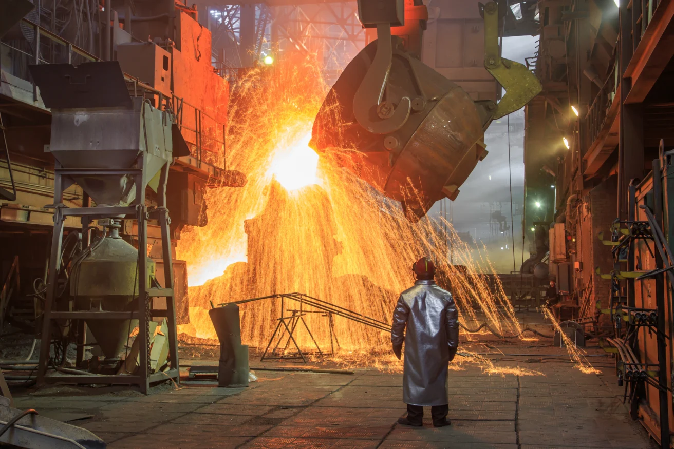 A worker in protective clothing stands near molten metal being poured from a large container, with sparks flying inside an industrial steel foundry.