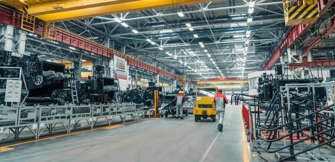 Interior of an industrial factory with machinery, workers in uniform, a yellow forklift, and large metal structures under bright overhead lighting.