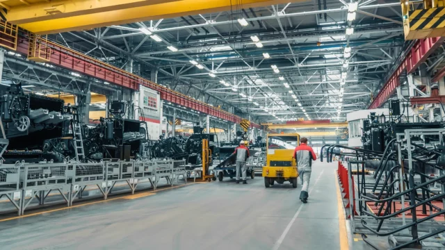 Interior of an industrial factory with machinery, workers in uniform, a yellow forklift, and large metal structures under bright overhead lighting.