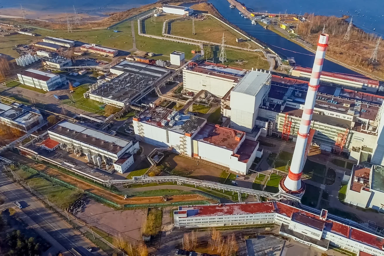 Aerial view of an industrial facility with multiple buildings, a tall striped chimney, roads, and waterways nearby.