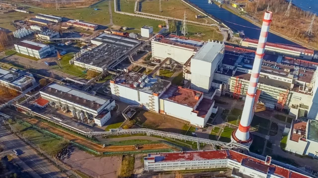 Aerial view of an industrial facility with multiple buildings, a tall striped chimney, roads, and waterways nearby.