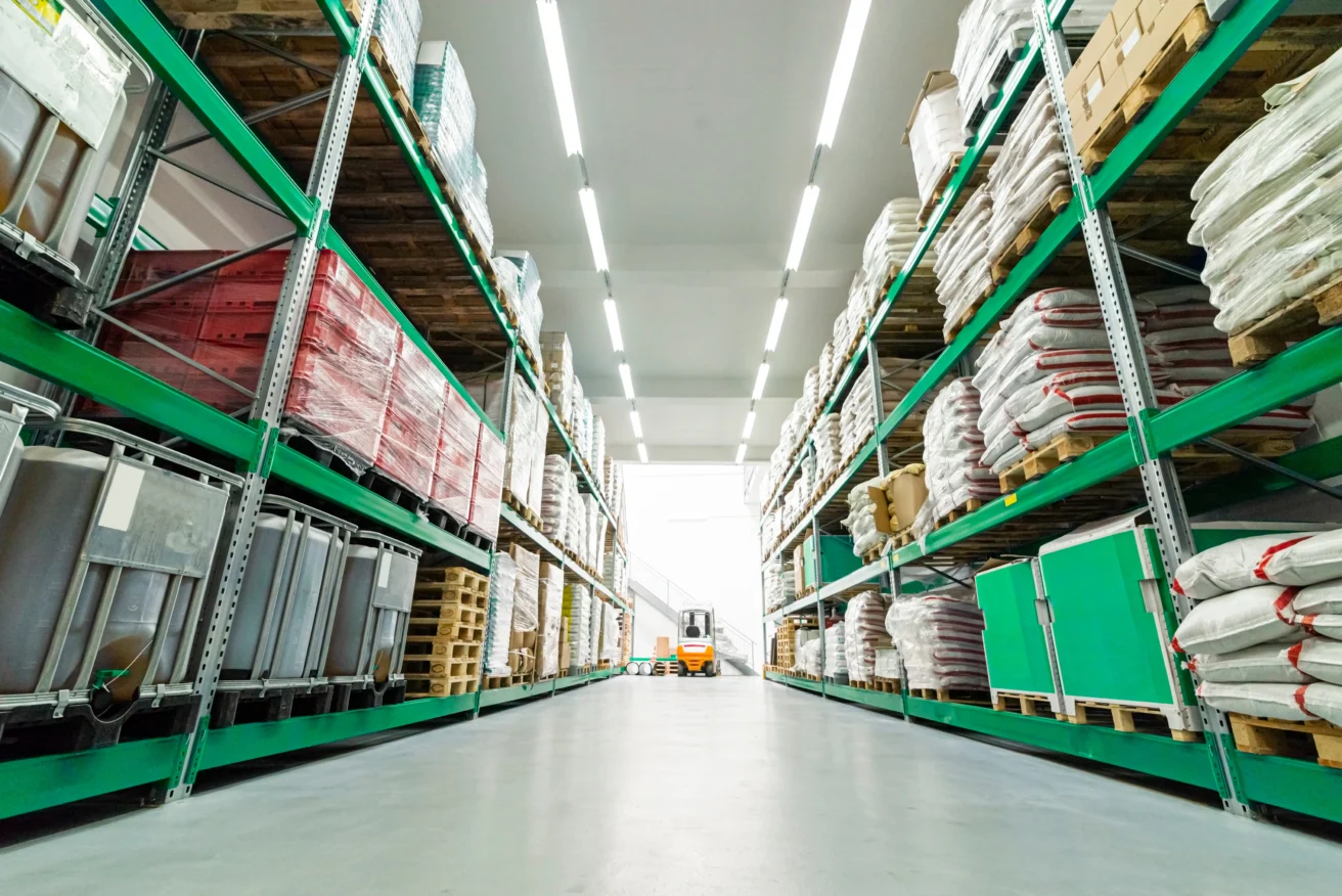 Warehouse interior with tall shelves storing various goods and materials, and a forklift visible at the end of the aisle under bright lighting.