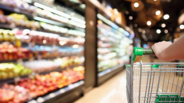 Person pushing a shopping cart down a grocery store aisle lined with produce on shelves.