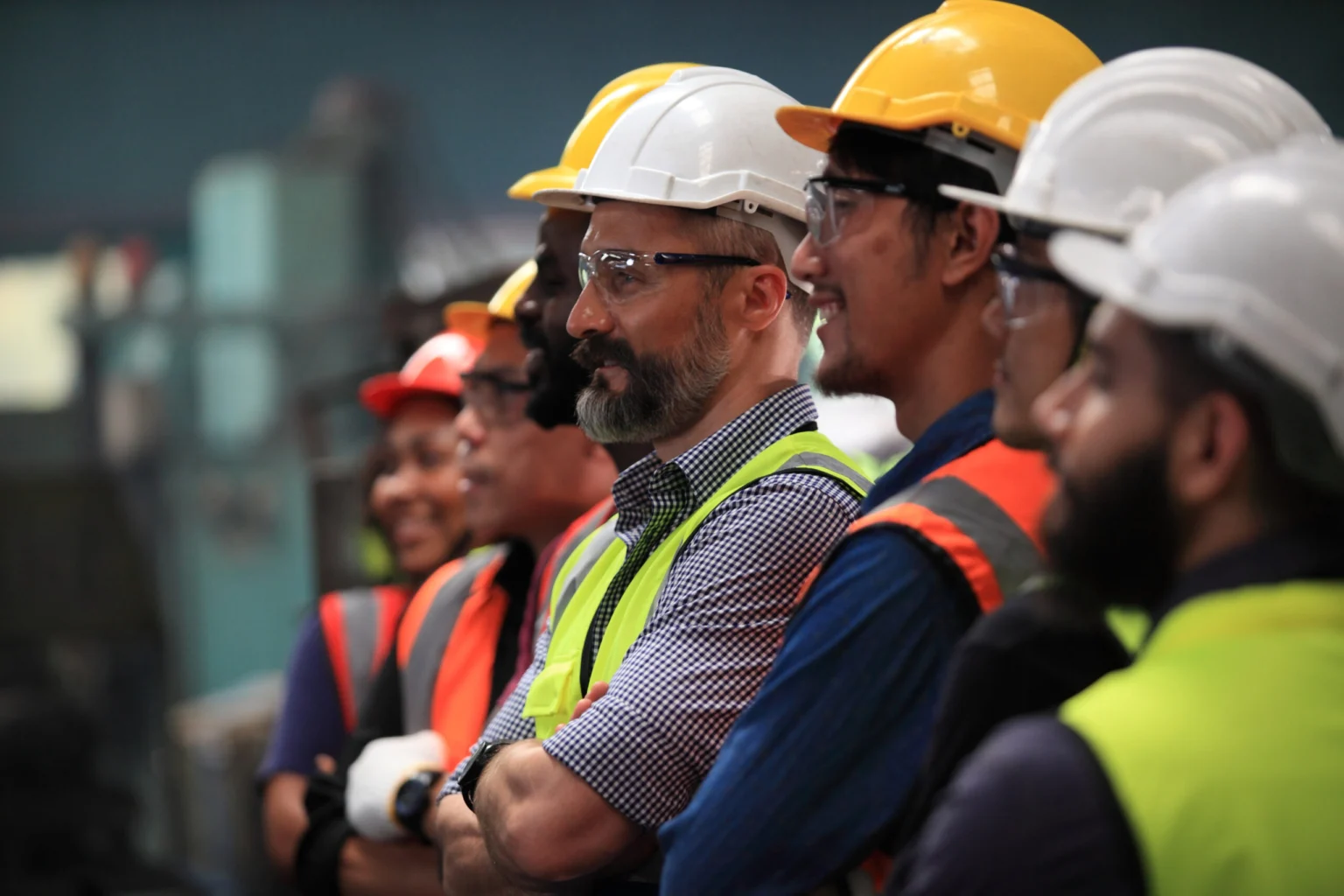 A group of construction workers wearing helmets and safety vests stands in a line, facing the same direction indoors.