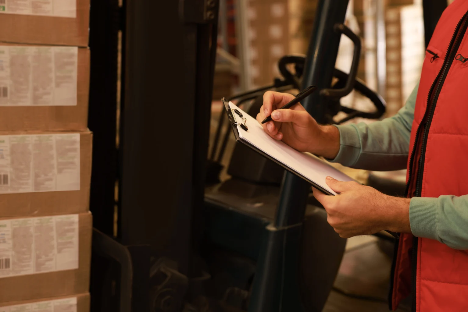 A person in a red vest writes on a clipboard near stacked cardboard boxes and a forklift in a warehouse setting.