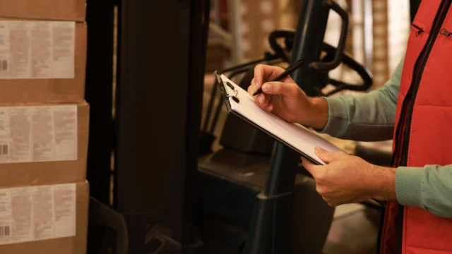 A person in a red vest writes on a clipboard near stacked cardboard boxes and a forklift in a warehouse setting.