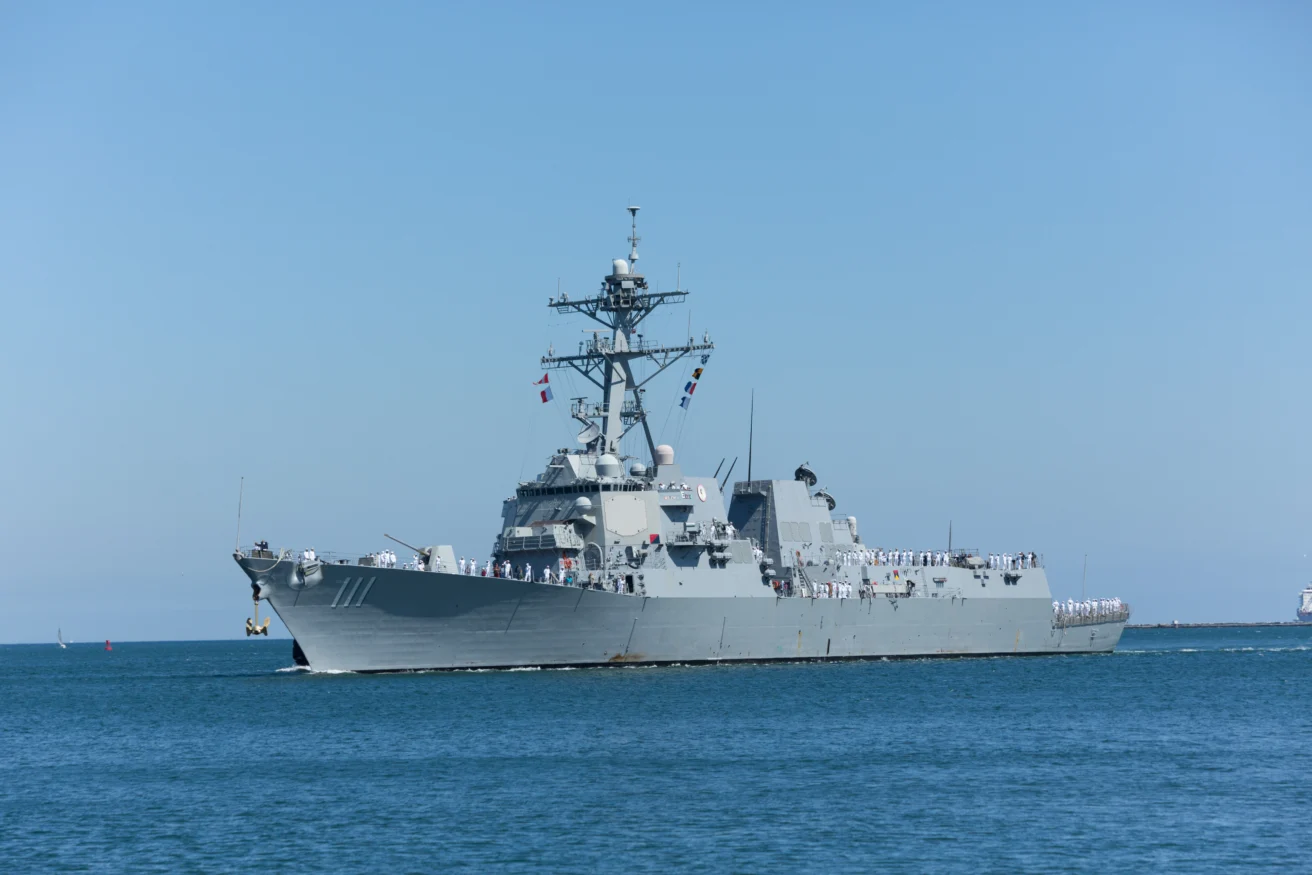 A large gray naval warship with crew members on deck sails on calm blue water under a clear sky.