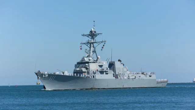 A large gray naval warship with crew members on deck sails on calm blue water under a clear sky.