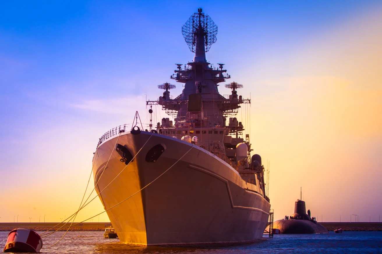 A large naval warship is docked at a harbor during sunset, with a submarine partially visible in the water beside it.