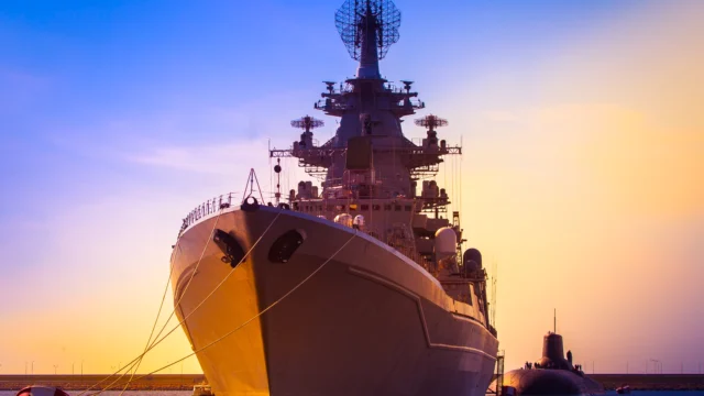 A large naval warship is docked at a harbor during sunset, with a submarine partially visible in the water beside it.