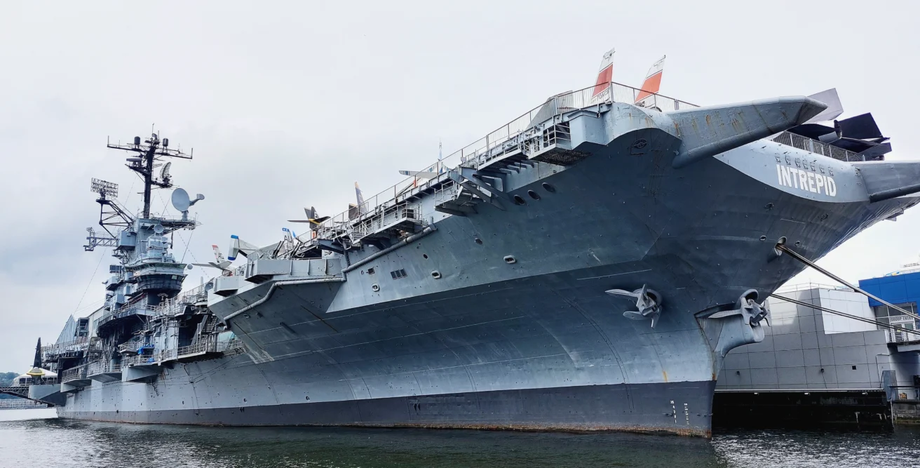 Large gray aircraft carrier named Intrepid docked at a pier, seen from a low angle, with flags and antennas visible on the upper deck.