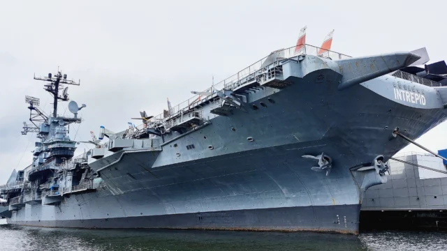 Large gray aircraft carrier named Intrepid docked at a pier, seen from a low angle, with flags and antennas visible on the upper deck.