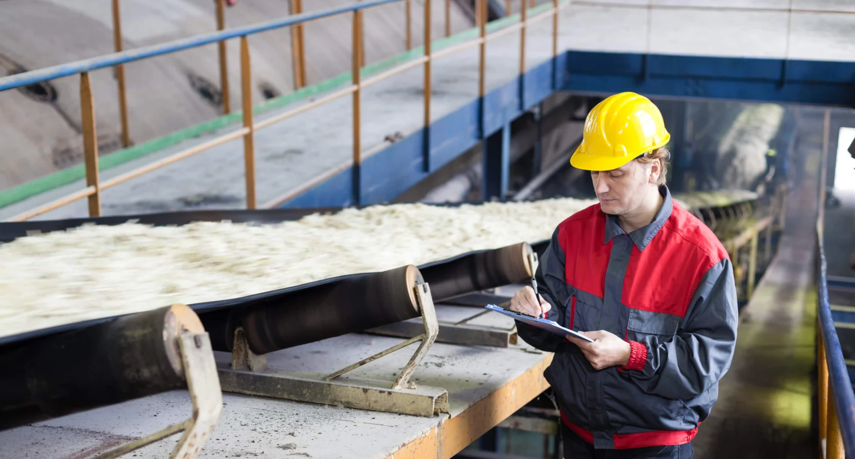 Worker in a red and gray uniform and yellow hard hat inspects a clipboard next to a conveyor belt transporting material in an industrial setting.