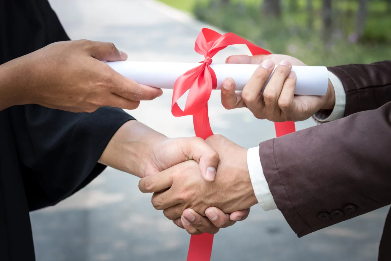 A graduate in a gown shakes hands with someone while receiving a diploma tied with a red ribbon.