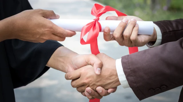 A graduate in a gown shakes hands with someone while receiving a diploma tied with a red ribbon.