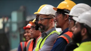 A group of workers wearing safety vests and hard hats stand side by side indoors, facing forward with attentive expressions.