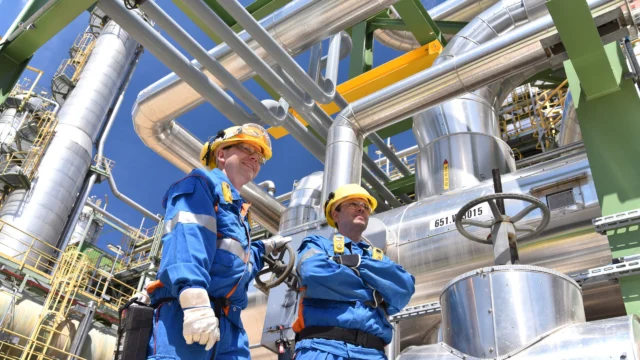 Two workers in blue uniforms and yellow helmets stand near industrial pipes and machinery at an outdoor facility, looking at the structures above them.