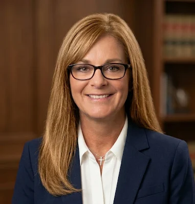 A woman with long light brown hair and glasses, wearing a navy blazer, white blouse, and cross necklace, smiles in an office setting with bookshelves in the background.