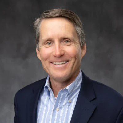 A person in a dark suit and striped shirt smiling at the camera against a gray background.