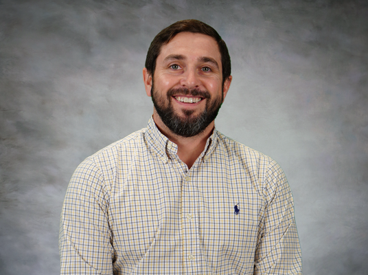 A person with a beard and short hair smiles, wearing a checked button-up shirt, against a gray background.